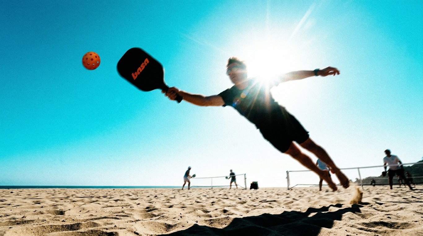 Player diving mid-air to strike a Basa pickleball on a sun-drenched beach court